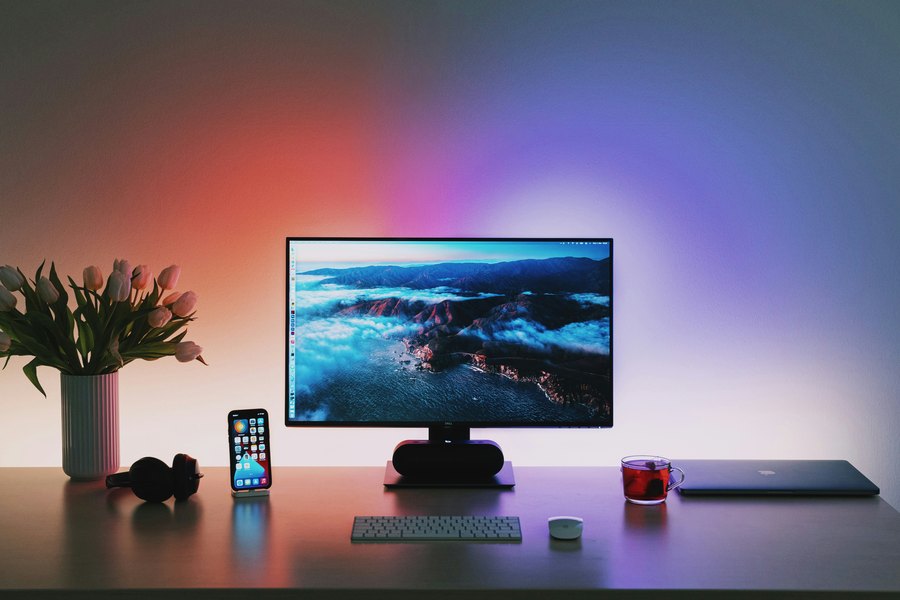 Schreibtisch mit Bildschirm, Tastatur und Laptop, farbiges Licht an der Wand